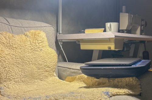 Nojatuoli pehmeällä lampaantaljalla, vieressä säädettävä lamppu ja pieni pöytä, jolla on teekuppi ja kirja. Kannettava tietokone odottaa käyttöä rauhallisessa kotiympäristössä.