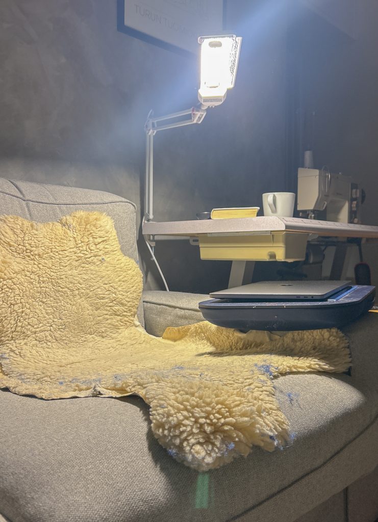 Nojatuoli pehmeällä lampaantaljalla, vieressä säädettävä lamppu ja pieni pöytä, jolla on teekuppi ja kirja. Kannettava tietokone odottaa käyttöä rauhallisessa kotiympäristössä.