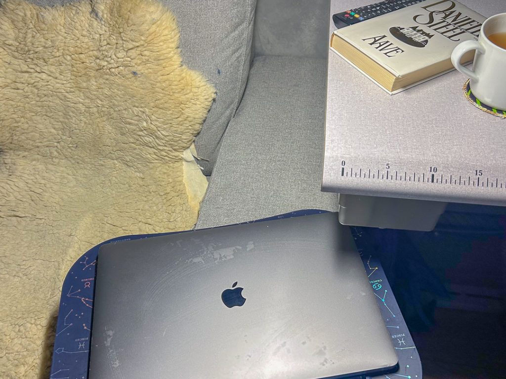 Sohvan vieressä pieni pöytä, jolla on teekuppi, kirja ja kaukosäädin. Edessä MacBook sylitasolla, ympärillä pehmeä viltti – rauhallinen koti- ja kirjoitushetki.
