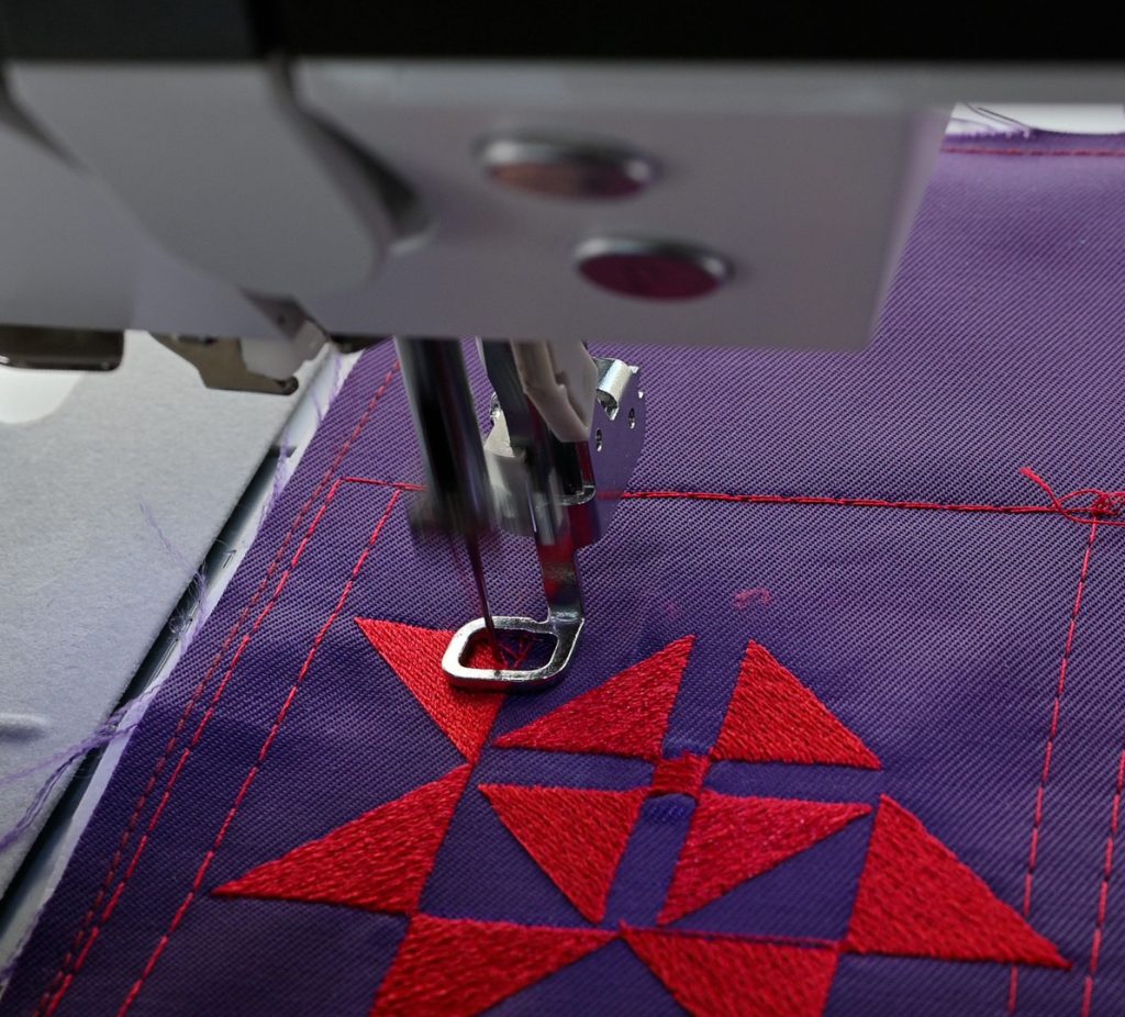 Kirjontakone tekee punaista geometrista kuviota violetille Enstex-kankaalle kirjontakehyksessä.