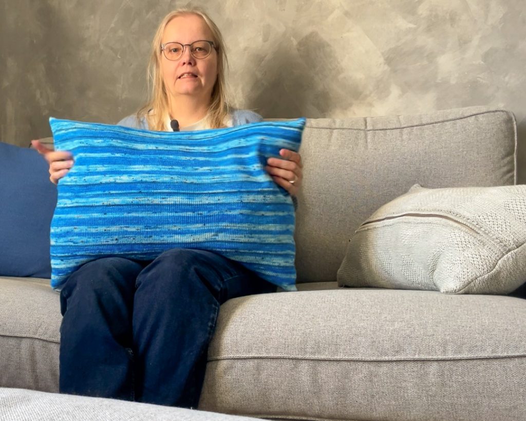 Heidi Salokangas esittelee suurta sinisävyistä raitatyynyä harmaalla sohvalla.