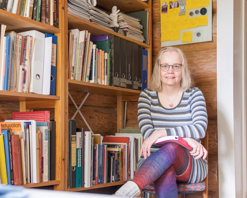 Heidi Salokangas istuu työhuoneessa yllään siniharmaavalkoinen raidallinen neulemekko.