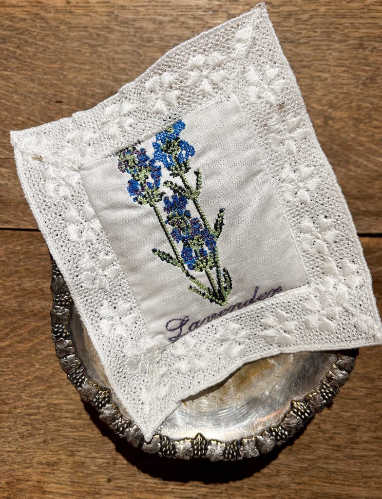 Valkoinen laventelityyny, jossa on sininen kukkakirjonta, Lavender-teksti ja leveä valkoinen pitsireuna.