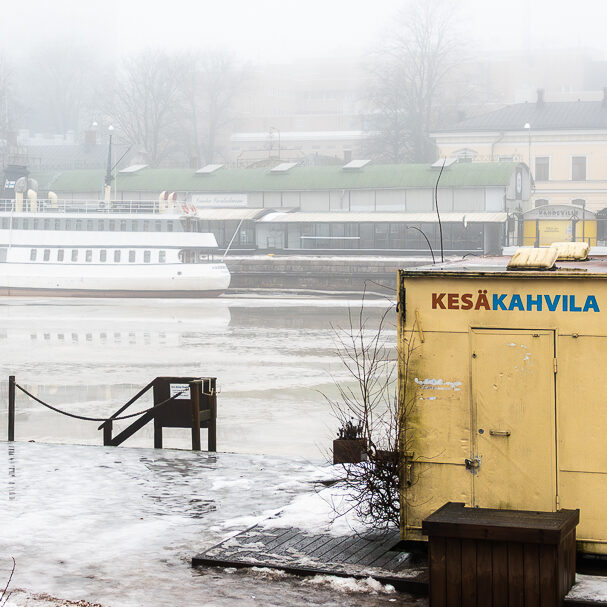 Sumuinen Aurajoki Turussa: jokiranta, vanha laiva ja keltainen kesäkahvilarakennus harmaassa talvisäässä.